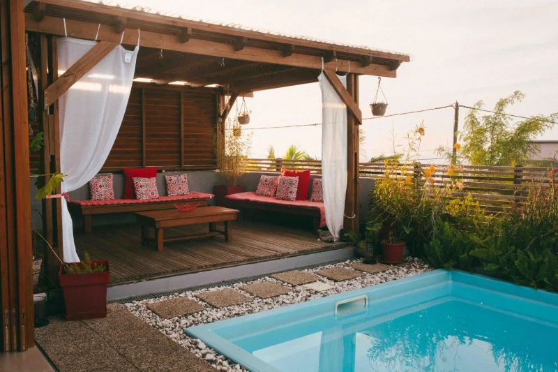 Le salon sous la pergola de la Kazanou, ses coussins rouges et la piscine, en fin de journée